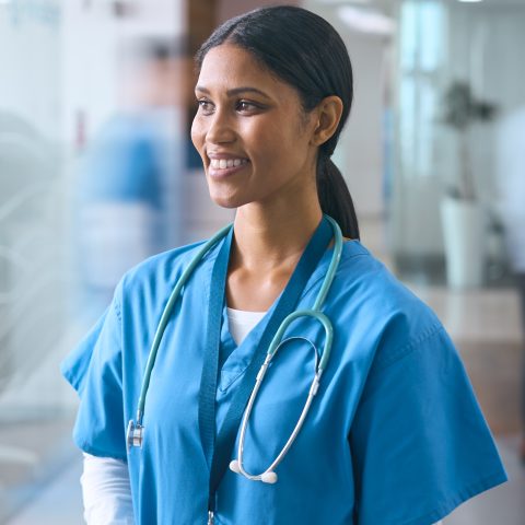 Female Doctor Or Nurse In Scrubs In Hospital Corridor With Colleagues Behind