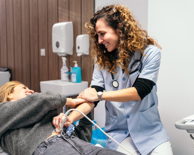 A female healthcare professional with curly hair is smiling reassuringly at a patient lying on an examination table while conducting an ultrasound scan.